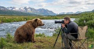 A photographer and a large grizzly bear sit facing each other at close range on a grassy riverbank with mountains in the distance.