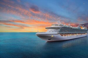 A large white multi-deck cruise ship sails from left to right on a calm blue ocean, heading slightly away from the viewer. The sky above features a gradient of deep blue transitioning to bright orange and yellow clouds on the horizon, suggesting sunrise or sunset. The ship's bow is prominent on the left, and its decks are illuminated with warm lights.