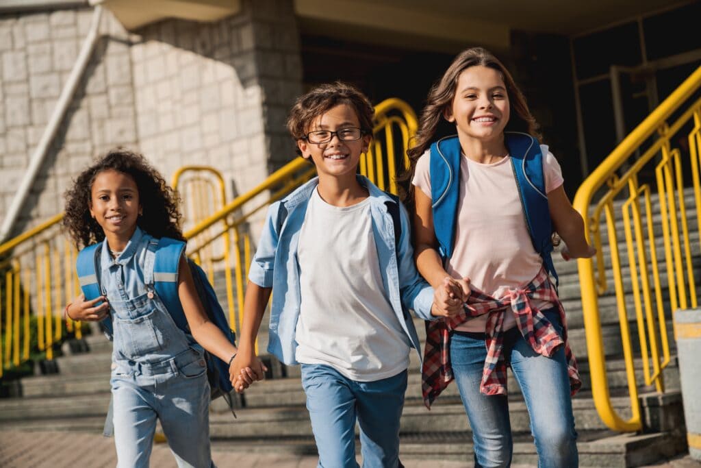 Happy kids elementary school pupils children with rucksacks backpack walking holding hands at school yard running out outside at the end of the lessons. Finish school and summer vacation concept.