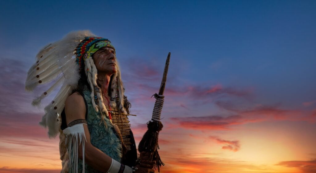 Indian red wearing traditional dress with bird feature on head holding weapon on hand with twilight sky background.