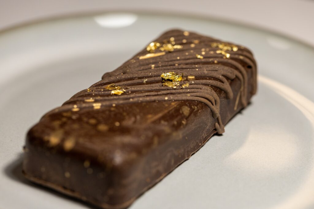 A premium Dubai chocolate bar coated in rich dark chocolate and topped with delicate edible gold flakes. The cut reveals a textured filling made with caramelized honeycomb and fine ingredients, emphasizing luxury and craftsmanship