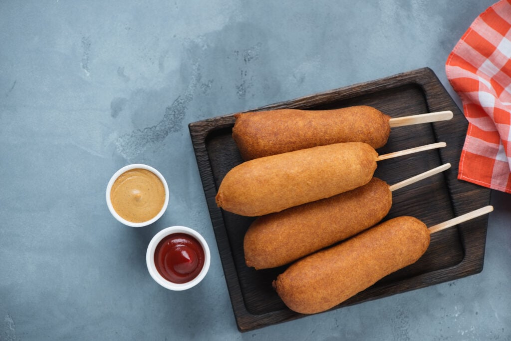 Corn dogs on a black wooden serving tray with mustard and ketchup. Flatlay with space over grey concrete background
