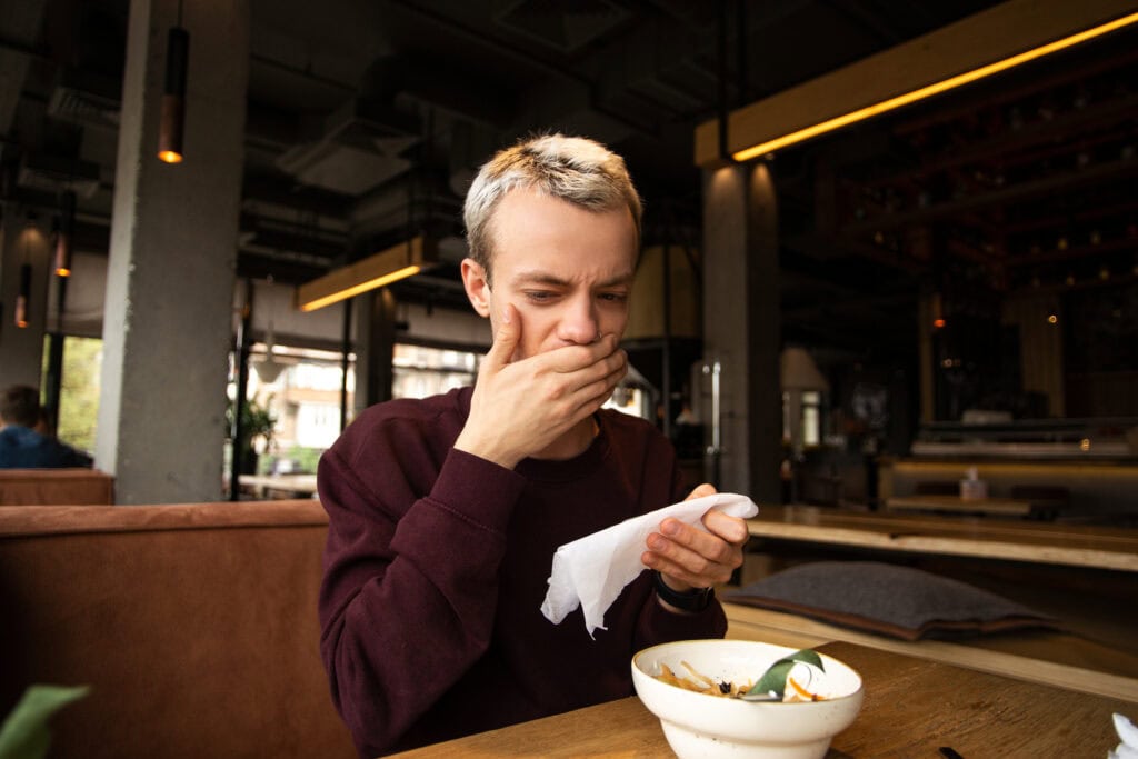 Spoiled food in the restaurant. Dissatisfied young man in a cafe covers his mouth with a hand disgusted with a taste of food. A meal on the table in front of him. Bad customer service concept.