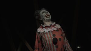Crazy creepy killer man dressed as clown with makeup at attic in ruined house looks at camera, talking, smiling going crazy. Halloween thriller concept.