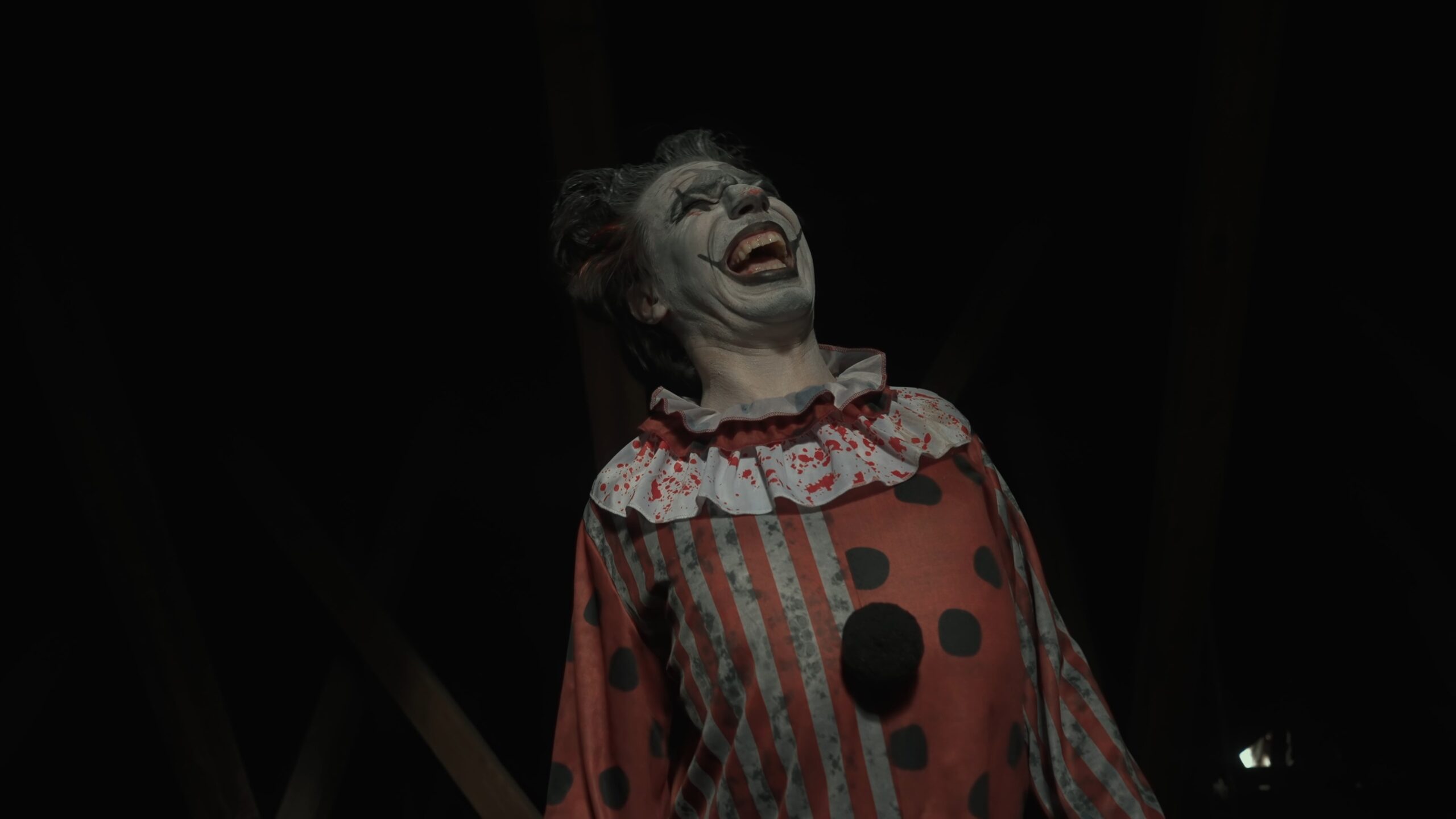 Crazy creepy killer man dressed as clown with makeup at attic in ruined house looks at camera, talking, smiling going crazy. Halloween thriller concept.