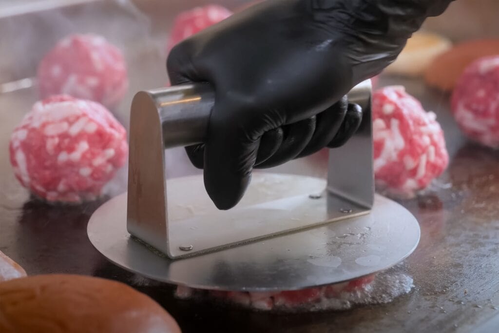 Cook uses a metal burger press to flatten raw ground beef balls on a hot griddle, with burger buns and steam in the background during commercial food prep

