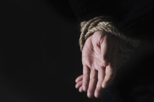 closeup of a man with his hands tied behind his back with rope, against a black background with some blank space on the left