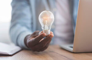 New idea concept. Unrecognizable black businessman holding illuminated light bulb in hand while sitting at workplace in office