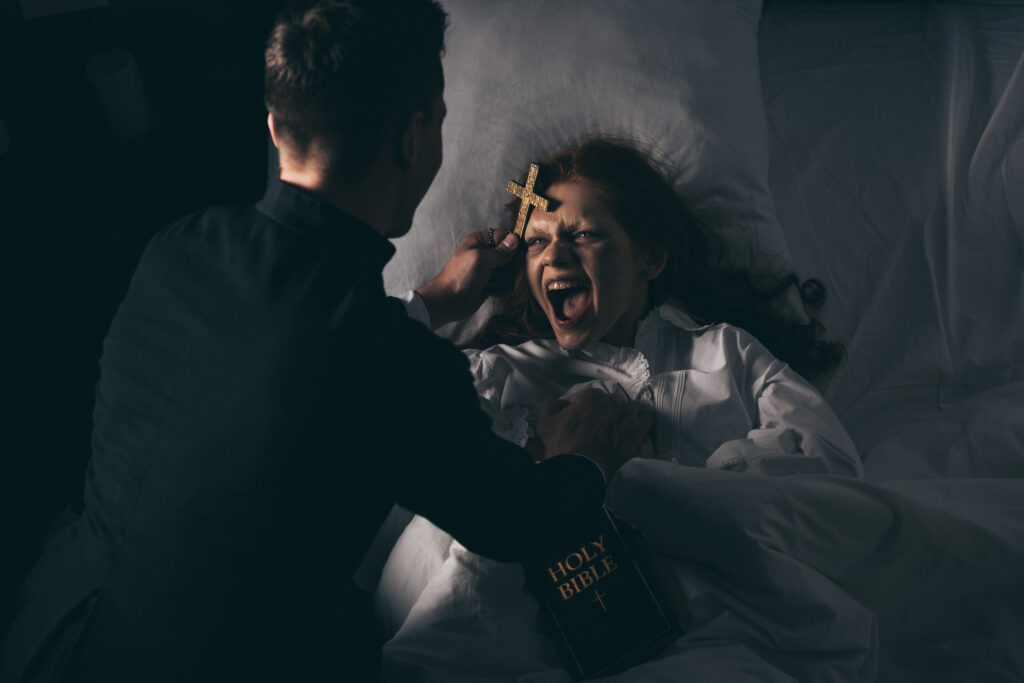 male exorcist with bible and cross standing over demoniacal screaming girl in bed