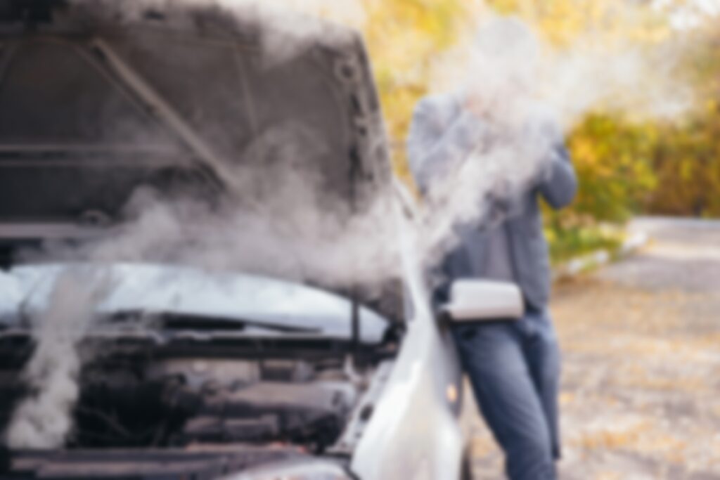 A man looks under the open hood of a car. The car broke down on the road. The engine is smoking. Blurred background