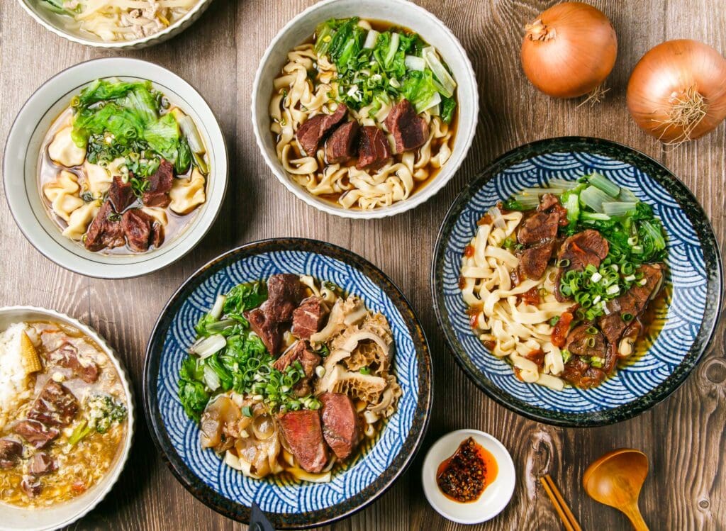 Assorted meat noodles Onion and tomato beef noodles, Sanbao noolde, dumplings soup with meat, Beef Cheeks, with chilli sauce, onion, wooden spoon, and chopsticks served side view of taiwanese food
