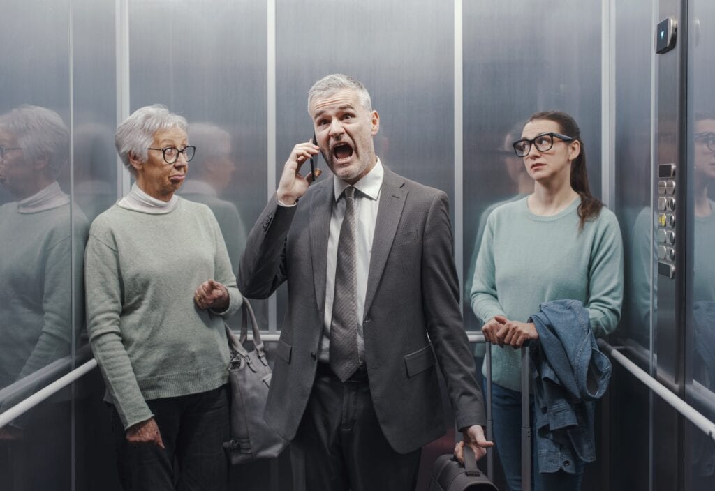Stressed angry businessman shouting into his smartphone in the elevator, two women are annoyed and looking at each other