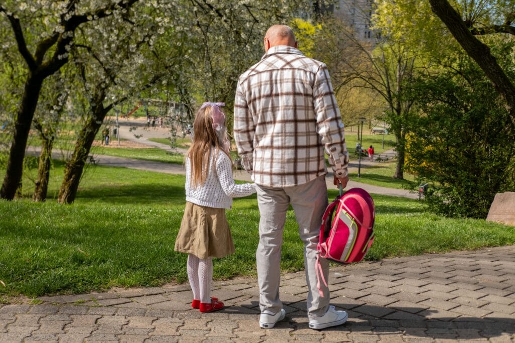 Grandfather walking hand in hand with granddaughter across sunny park, little girl wearing a pink school backpack, Walking to school spring day, education concept, family bonding, outdoor lifestyle