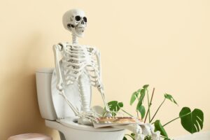 Human skeleton with magazine sitting on toilet bowl in restroom
