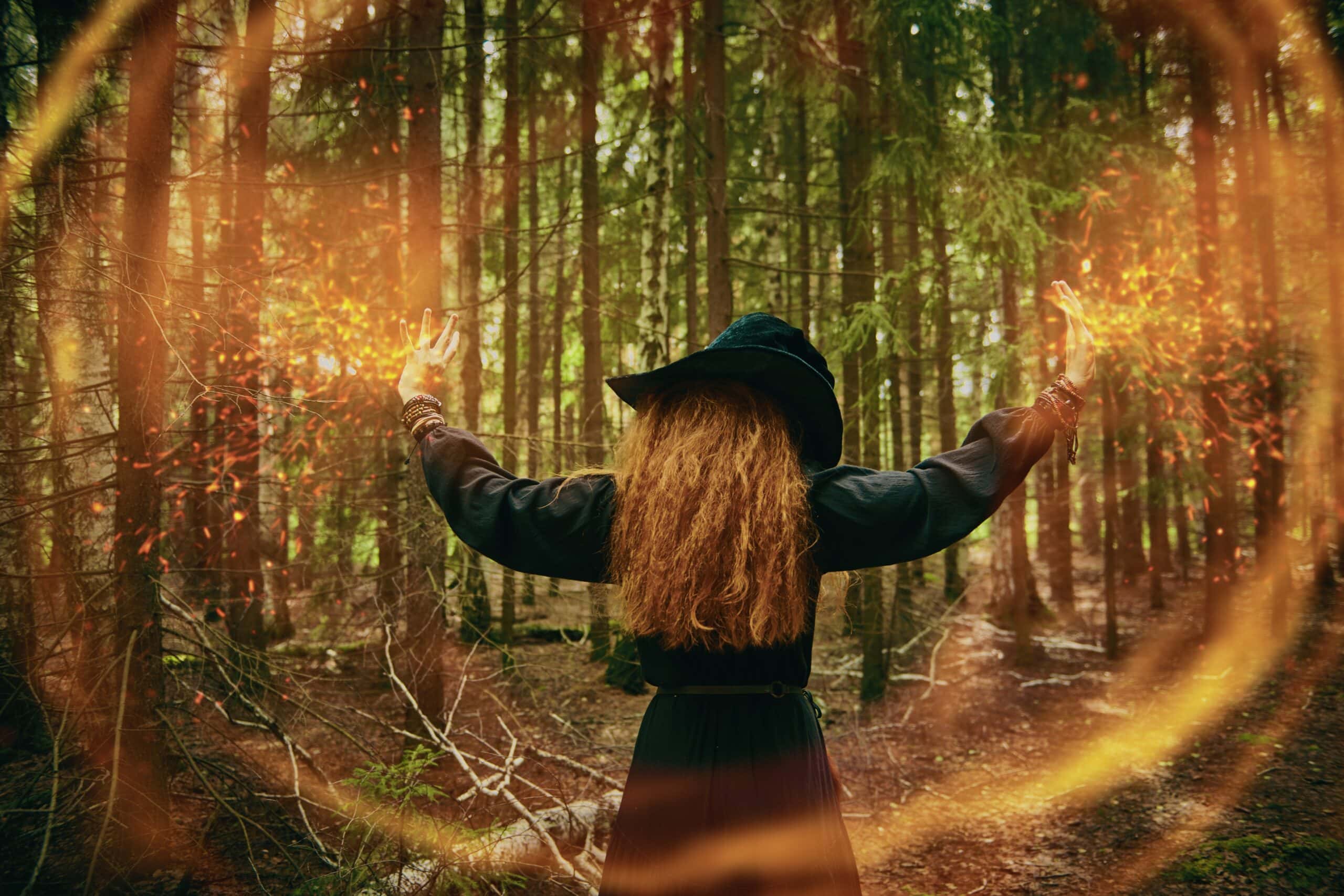 A powerful forest witch with red hair, dressed in a black dress and a large black hat, casts a spell, in a dense forest. Copy Space. Halloween.