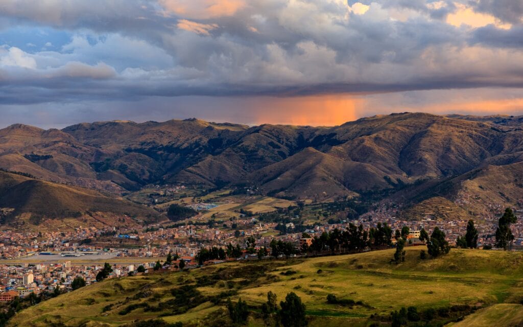 Scenic view of Cusco, Peru, with lush hills and dramatic clouds during sunset, showcasing the city's unique landscape.