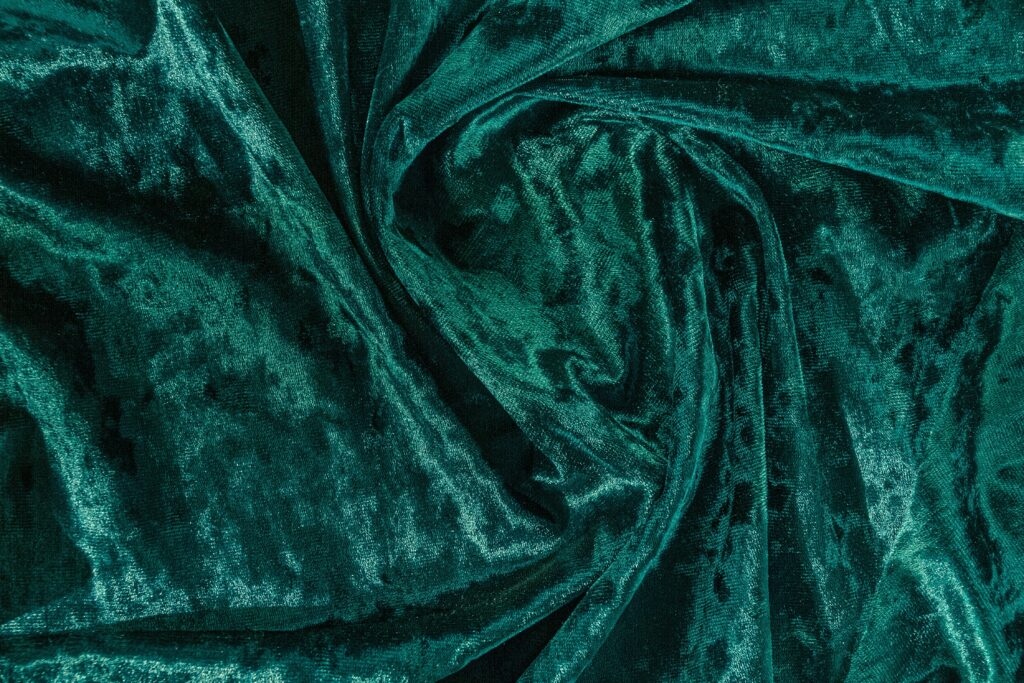 Top-down, close-up view of luxurious dark teal or emerald green crushed velvet fabric, featuring deep folds, plush texture, and shimmering highlights