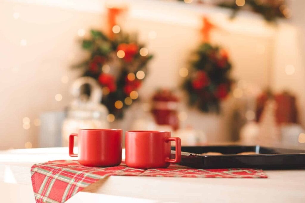 Two red ceramic cup of coffee with baked biscuits on metal baking tray on kitchen table over Christmas decorations with lights close up. Winter holiday season. Good morning.