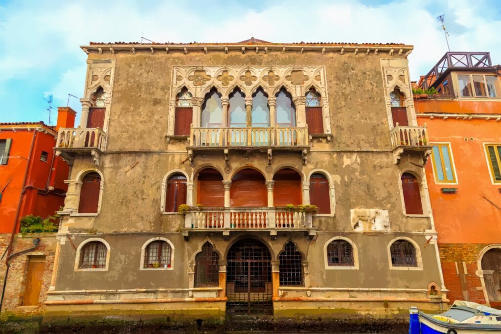 Venice, Italy. A ground-level view of the Ca' Mastelli, also known as the Palazzo Mastelli del Cammello or the Camel House, a Venetian Gothic palace fronting a canal. The three-story facade features a weathered ochre stucco exterior with white Istrian stone detailing. A grand, ornate four-light window with intricate stone tracery dominates the first floor, opening onto a small wrought-iron balcony. Below, an arched water portal with a double wooden door is flanked by tall, narrow windows with dark shutters. A small statue is positioned to the right of the entrance. The greenish-brown canal water reflects the cloudy sky and surrounding architecture, with a blue-and-white boat moored nearby. Built by merchant brothers in the 12th century, the palace gets its popular name from a bas-relief of a man leading a camel on its facade, a sculpture tied to a legend of a lovelorn merchant.