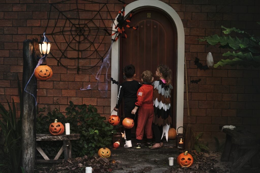 Three children in Halloween costumes trick-or-treating at a door. Pumpkins and cobwebs decorate the scene. Halloween fun with costumes and pumpkins. Halloween celebration image.