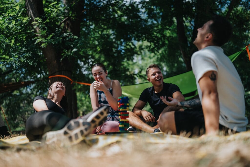 A group of friends relax on a blanket in a green park, laugh while stacking colorful blocks in a playful outdoor game. Casual summer hangout with smiles and friendship.