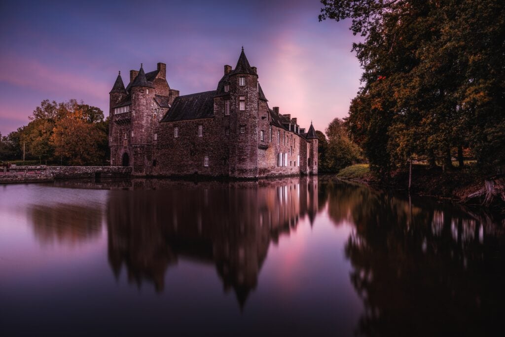 The Château de Trécesson stands like a timeless medieval fortress nestled on the edge of a tranquil lake in the heart of Brittany’s legendary Brocéliande Forest. Its deep reddish‑schist walls rise with a striking presence, mirroring perfectly in the calm water that surrounds the castle’s moat. A stone bridge leads across the moat to an imposing gatehouse flanked by narrow towers and connected by a historic gallery. The steep slate roofs and fortified structure retain an authentic 15th‑century character, evocative of centuries past. Set against dense forest and open sky, Trécesson blends architectural strength with natural serenity, offering a poetic view of France’s rich heritage and romantic medieval landscapes