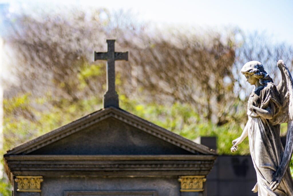 Statue of an angel beside a tomb with a cross on top. The blurred background creates a soft contrast between the sacred and nature. Artist unknown, São João Batista Cemetery, Rio de Janeiro, Brazil.